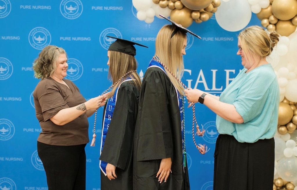 Two graduates in black caps and gowns stand side by side while two people place honor cords around their necks in front of a blue “Pure Nursing” backdrop decorated with white and gold balloons.