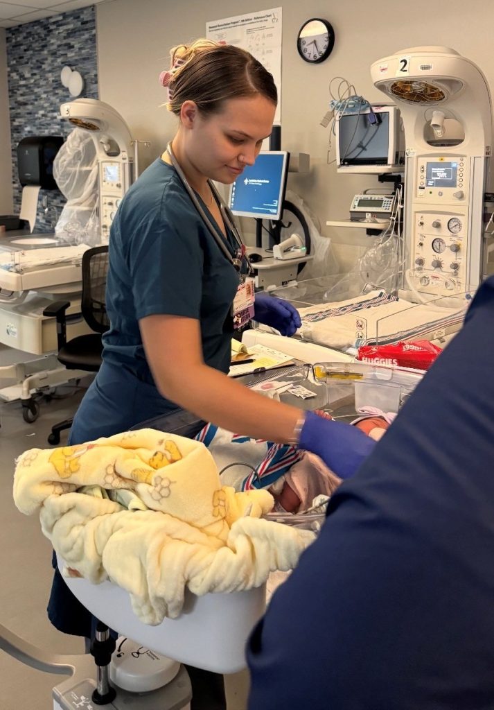 A nursing student in scrubs and gloves works at a hospital bassinet, caring for a newborn in a clinical room with medical equipment, monitors, and infant warmers visible in the background.
