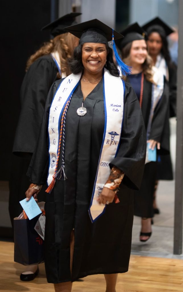 A nursing graduate wearing a cap, gown, and nursing stole walks in a graduation procession while holding program materials.