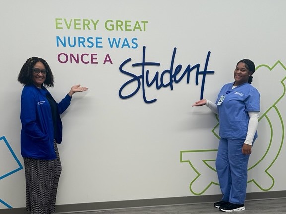 Two individuals pose in a hallway, standing in front of a wall that reads “Every great nurse was once a student,” with an illuminated exit sign above them.