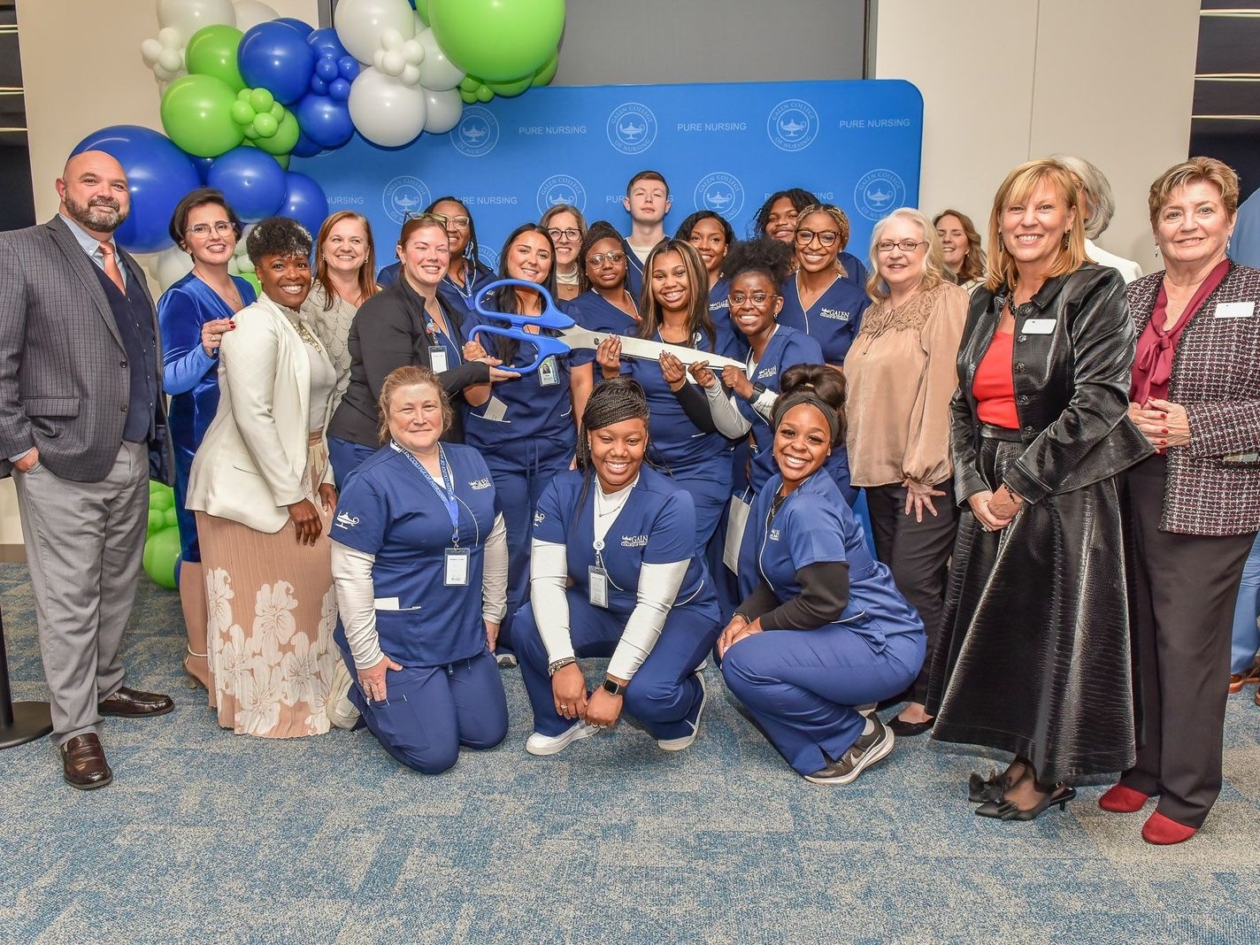 Large group of nursing students in blue scrubs posing with faculty and staff at a ribbon‑cutting event with balloons and a “Pure Nursing” backdrop.
