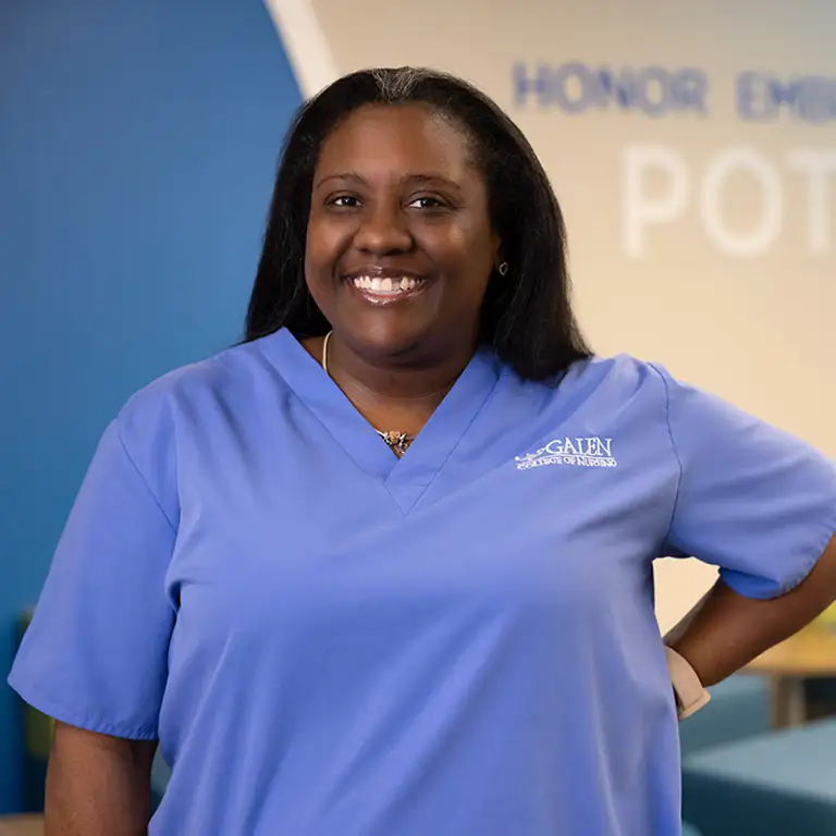A smiling woman with dark hair, wearing a light blue scrub top with a 'GALEN COLLEGE OF NURSING' logo, poses with one hand on her hip. She has silver strands in her hair and a necklace. In the blurred background, there's a blue and tan wall with large, light-colored text that reads 'HONOR EMB[RACE] POT[ENTIAL]'.