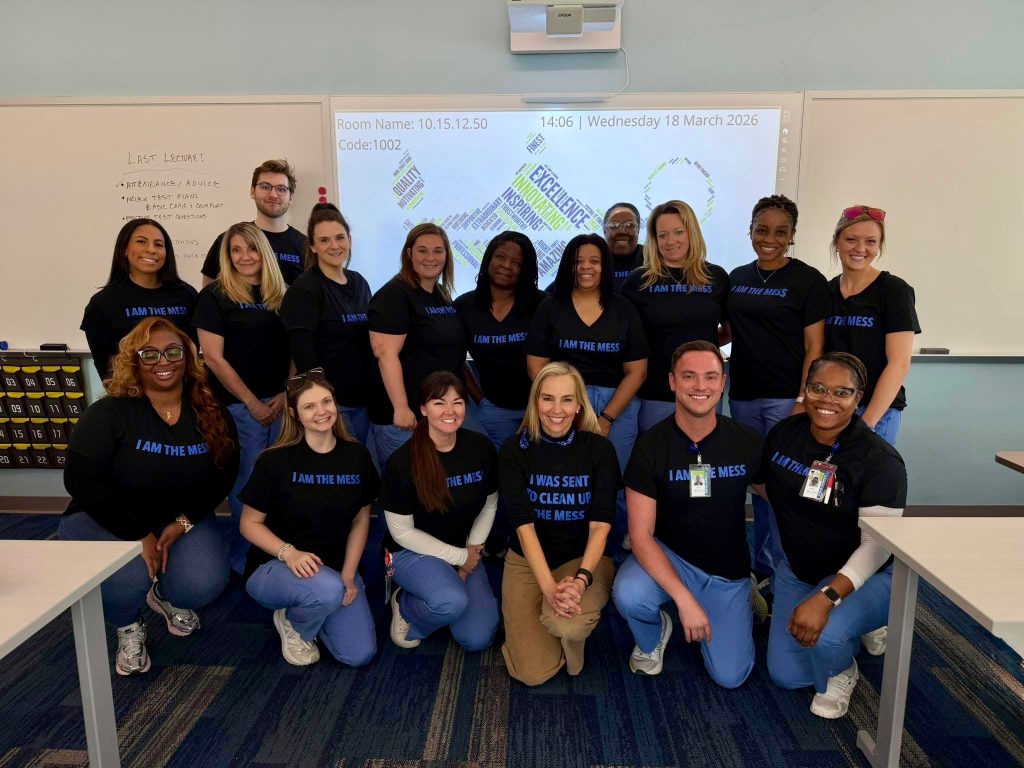 A group of Galen College of Nursing students and faculty stand together in a classroom wearing matching shirts, with a presentation screen behind them.
