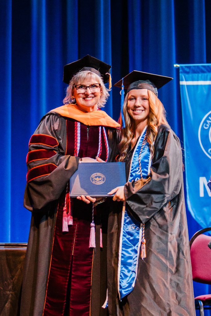 Graduate receives diploma on stage while standing beside faculty member.