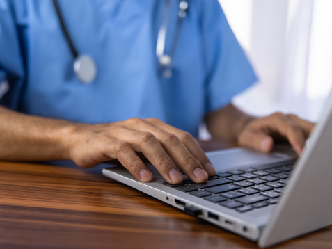 Nurse in blue scrubs typing on a laptop.