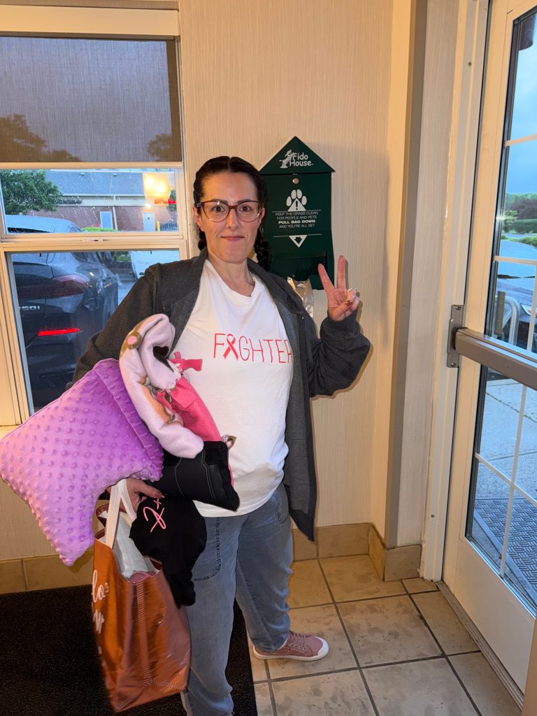 Person standing indoors near a doorway holds a folded blanket and makes a peace sign while wearing a white shirt with the word “Fighter” and a pink breast cancer ribbon. A window with rain visible outside and a wall-mounted pet waste bag dispenser are in the background.