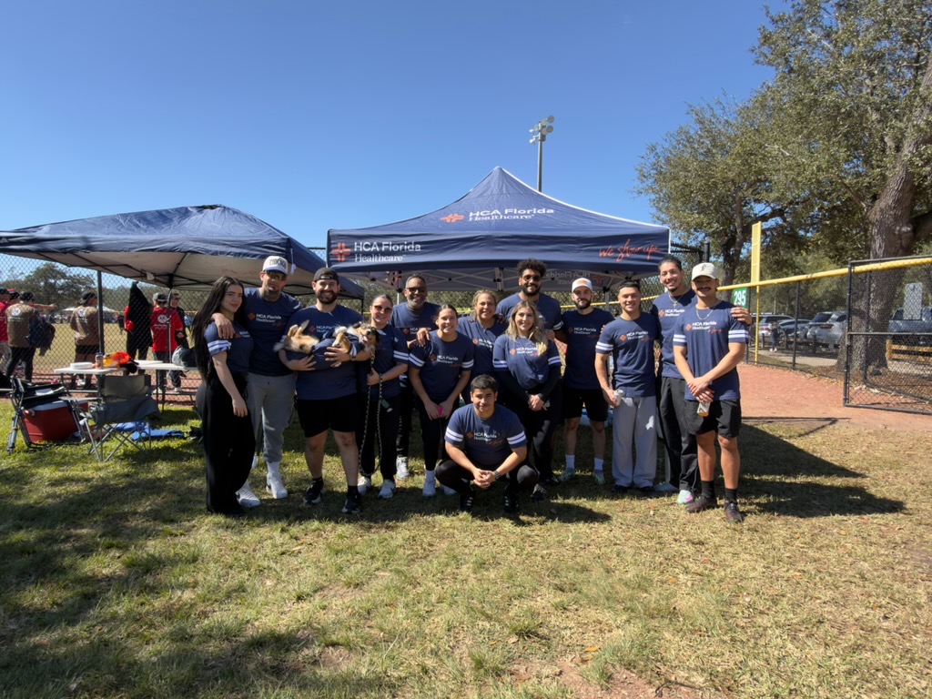 Group of participants standing together outdoors in front of an HCA Florida tent at a community event.