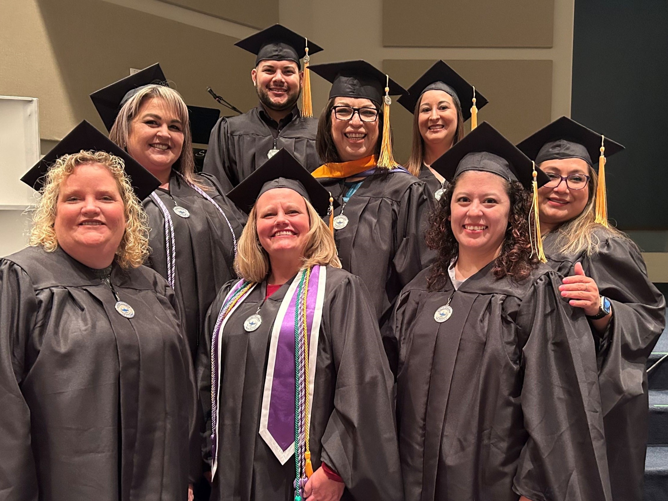 A group of graduates wearing black caps and gowns stand together indoors on a stage or platform. Several individuals have honor cords and stoles draped over their gowns, indicating academic achievements. The background shows neutral-colored walls and steps leading up to the group.