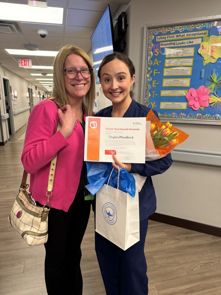 Two adults pose together in a hospital hallway holding a Flower Bud Award certificate and gift bag.