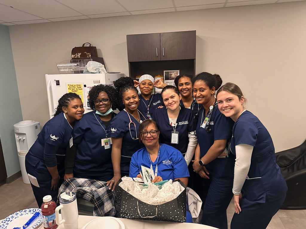 A group of nursing professionals in scrubs stand together in a break room, gathered around a seated colleague holding a gift bag.