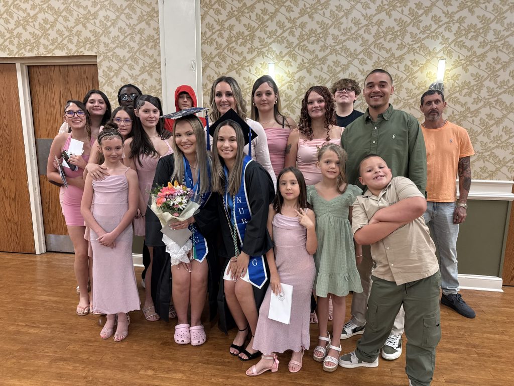 Two graduates in caps and gowns pose with a large group of family members and friends indoors, with one graduate holding a bouquet while others stand closely together behind and beside them.