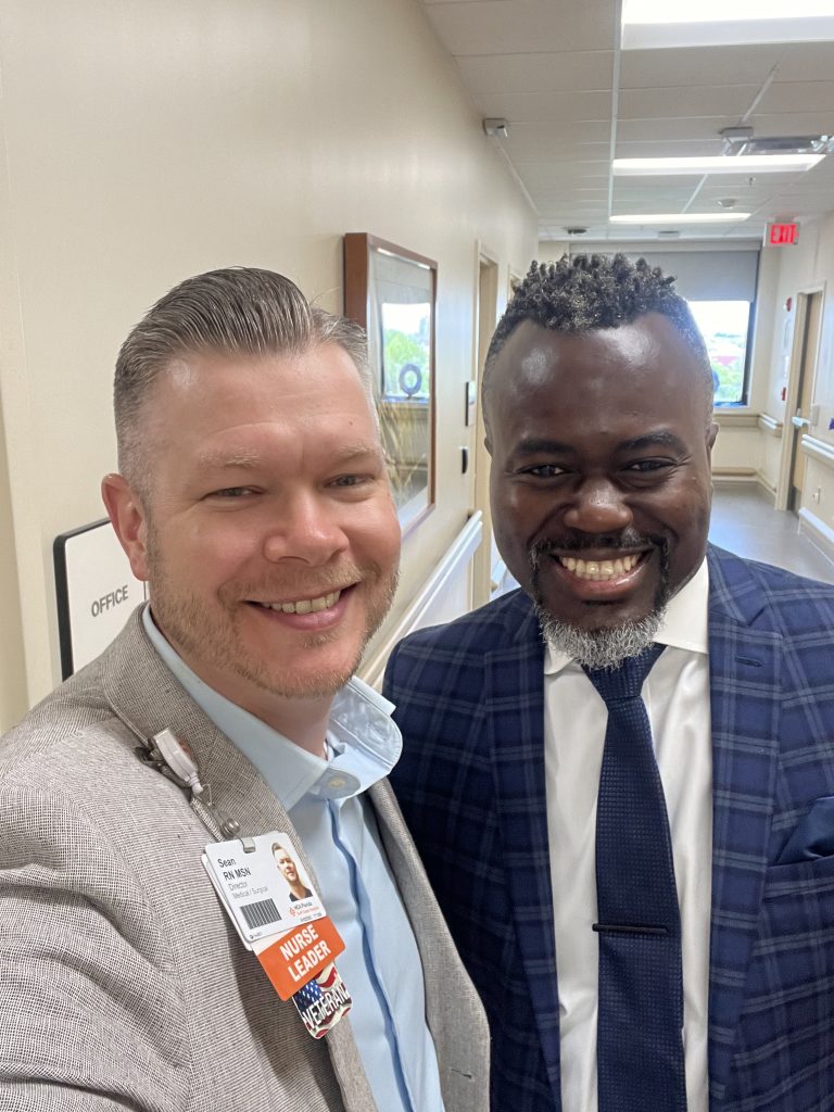 Two healthcare leaders standing together in a hospital hallway wearing professional attire and ID badges.