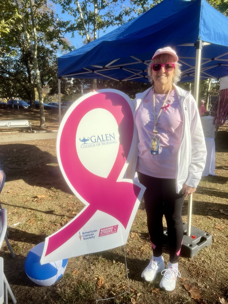 A person standing next to a large pink ribbon-shaped sign with the Galen College of Nursing logo and the American Cancer Society branding. The scene is outdoors under a blue canopy tent, with autumn leaves scattered on the ground.