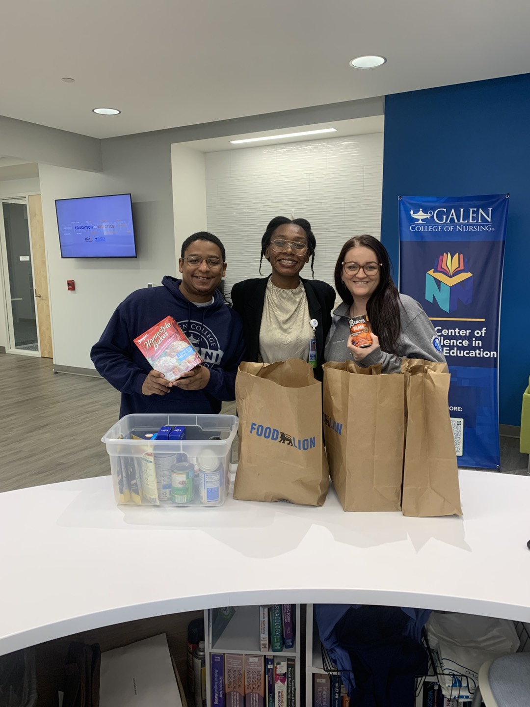 Three people stand with food items behind baskets from a local grocery store at Galen College of Nursing's Roanoke campus.