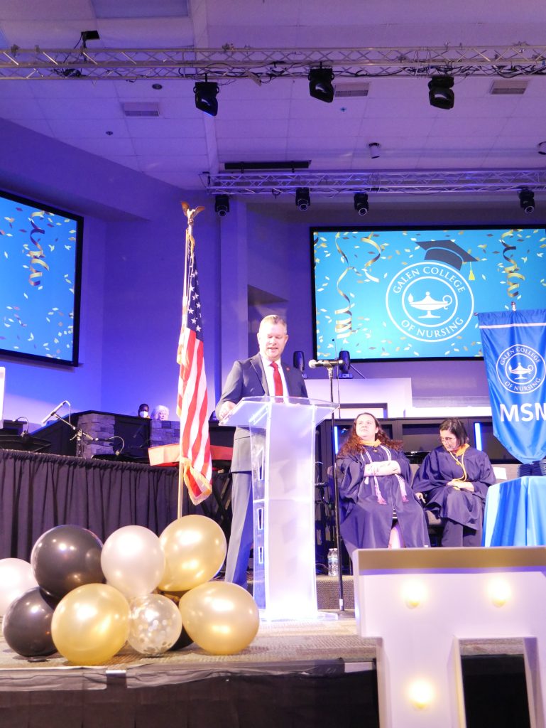 Speaker standing at a podium on stage during a Galen College of Nursing graduation ceremony with faculty seated behind.