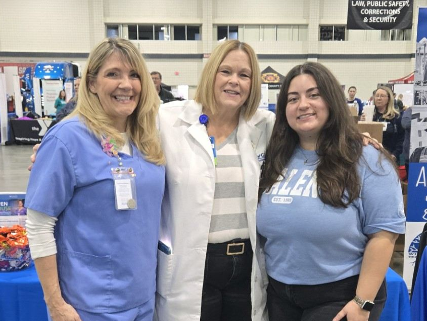 Three Galen College of Nursing representatives stand in front of a Galen table at a community event, with nursing education materials and medical training mannequins visible in the background.