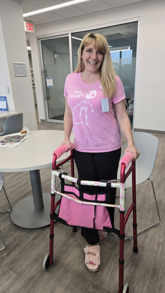A Galen College of Nursing student stands indoors using a walker, wearing a Galen badge, with campus meeting space furniture visible nearby.