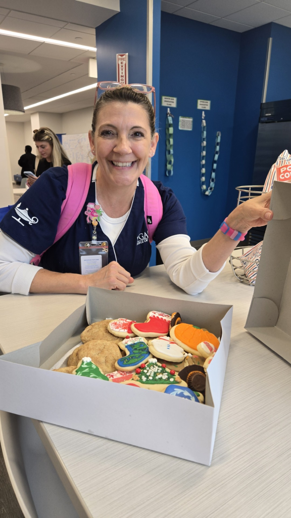 A Galen College of Nursing representative sits at a desk holding a box of decorated cookies, with campus signage and materials visible in the background.