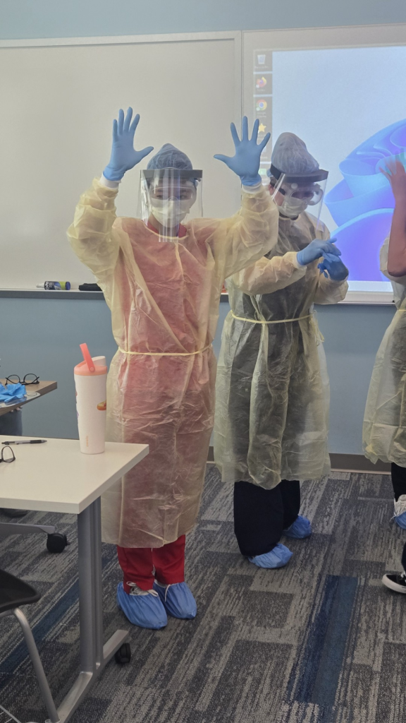 Two students in full protective equipment raise gloved hands while standing in a classroom, with other similarly dressed classmates nearby.