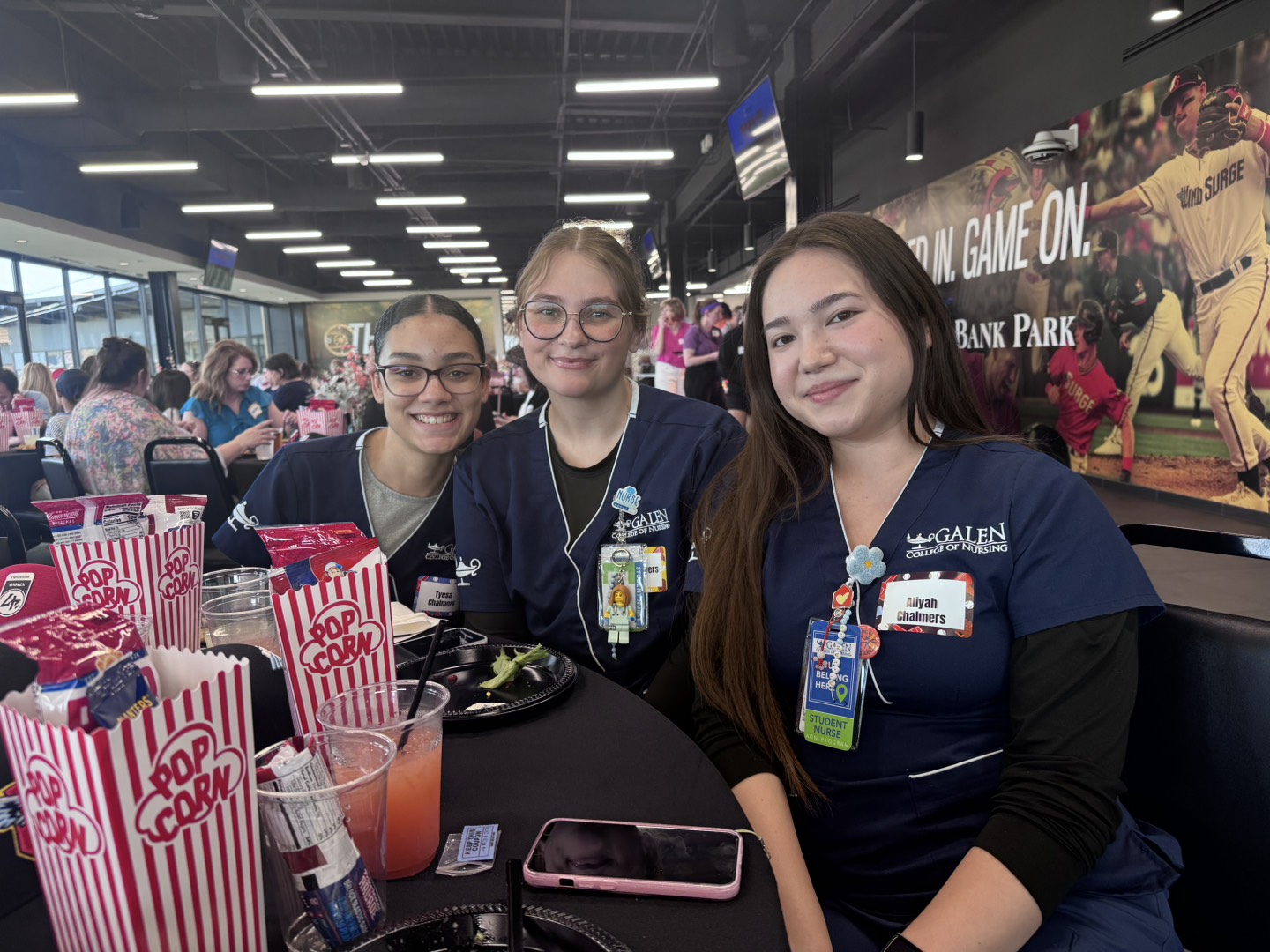 Three nursing students seated at a table with popcorn and drinks, smiling toward the camera in a busy event space with murals and attendees behind them.