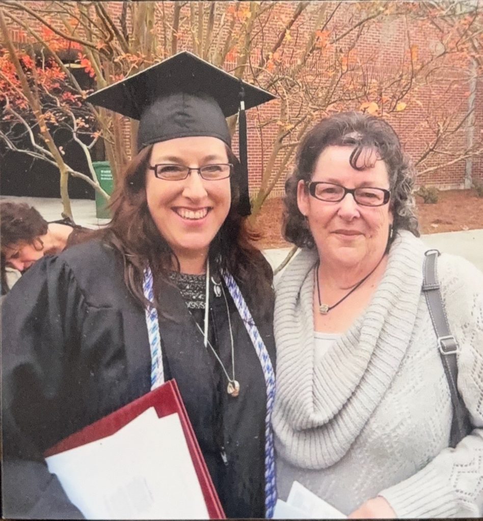 Graduate wearing a cap and gown holds a diploma folder while standing outdoors beside another adult near a brick campus building.