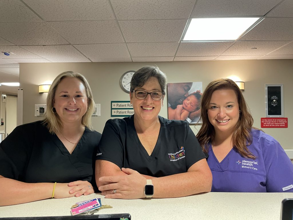 Three clinical staff members stand behind a hospital nurses’ station inside a patient care unit.