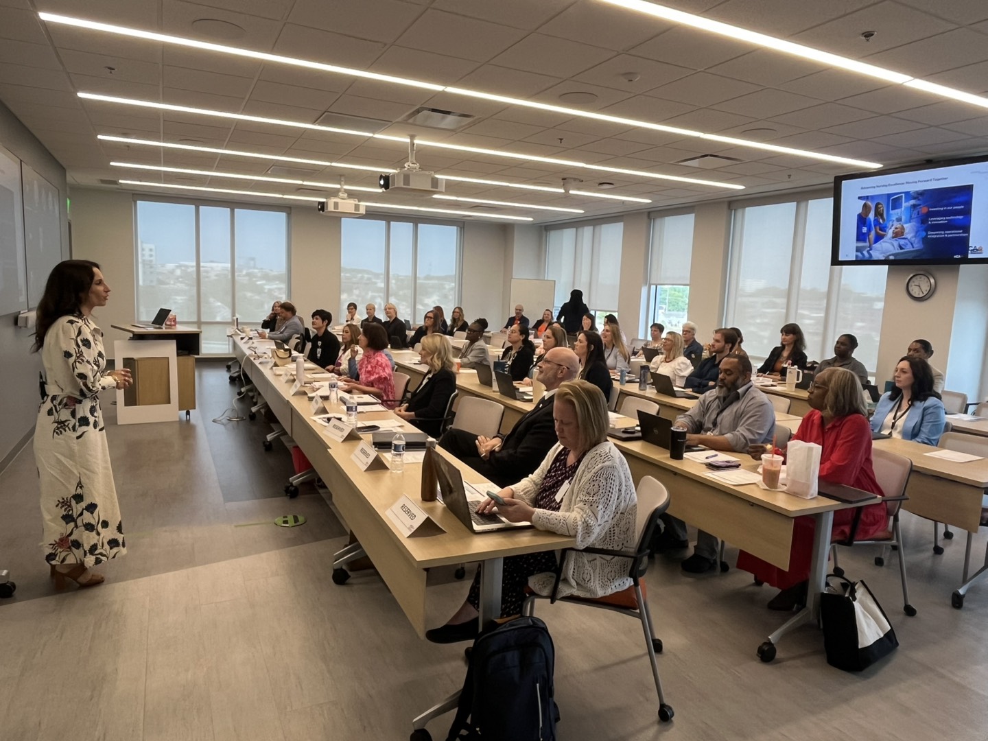 A classroom-style session with attendees seated at desks using laptops, listening to a speaker at the front near a presentation screen showing nursing education content.