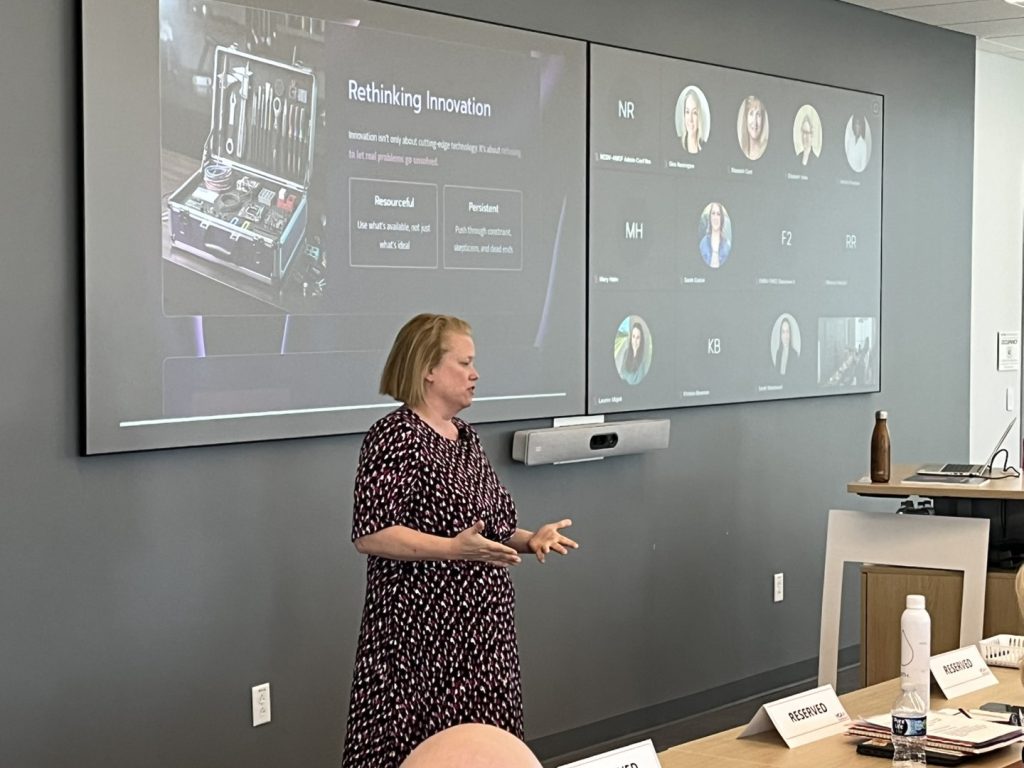 Speaker presenting a slide titled ‘Rethinking Innovation’ in a meeting room with a hybrid audience displayed on a large screen.