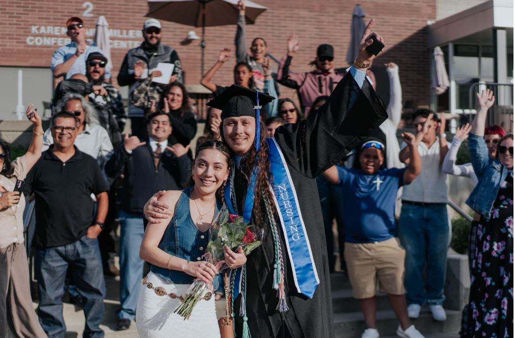A graduate wearing a black cap and gown with a blue “Nursing” stole stands with an arm around another person holding a bouquet of red roses. The graduate raises one hand while a cheering crowd behind them celebrates outside a brick building.