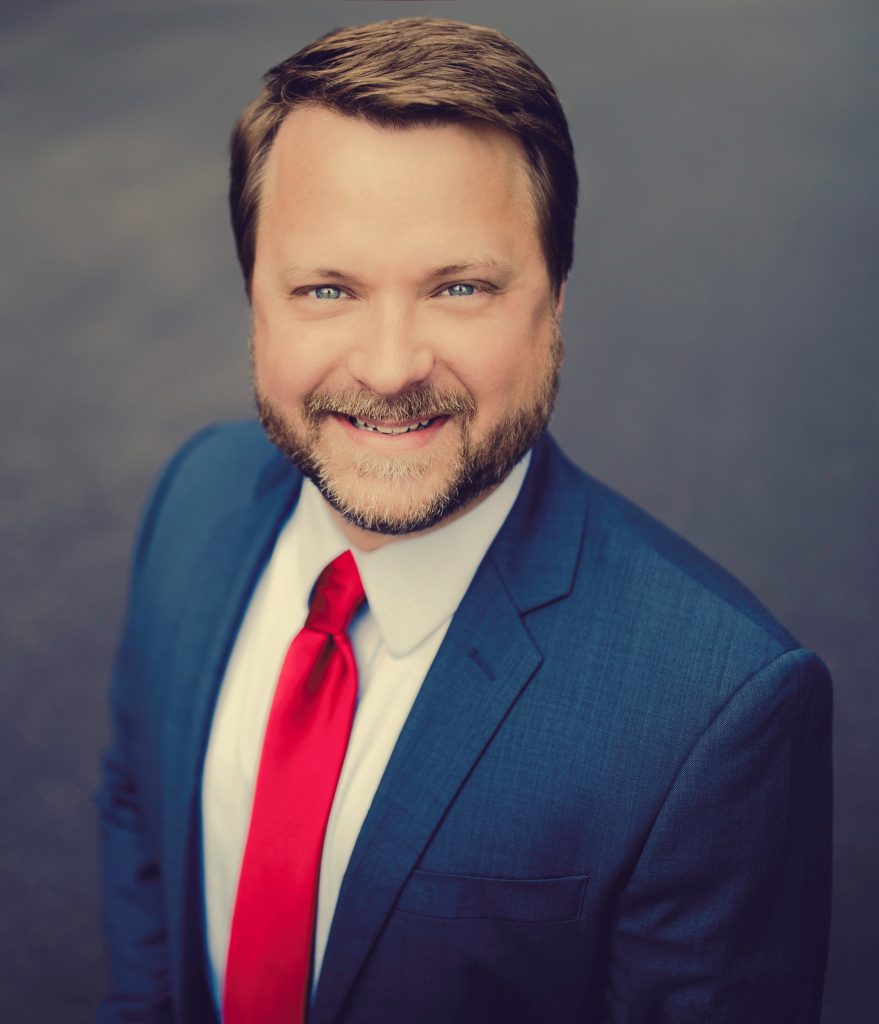 Professional headshot of a man wearing a navy suit, white shirt, and red tie.