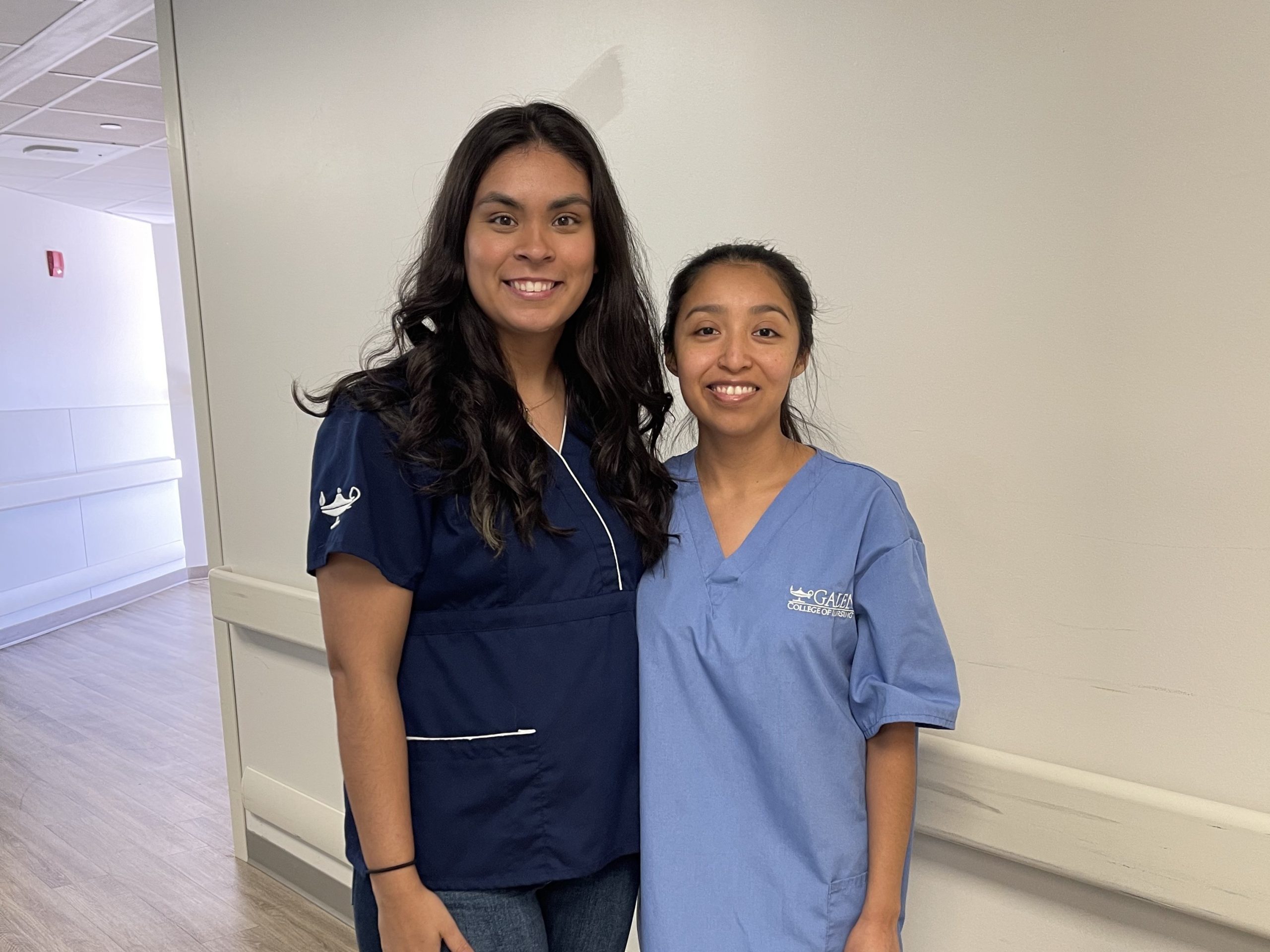 Two women smile for a photo in front of a white wall.