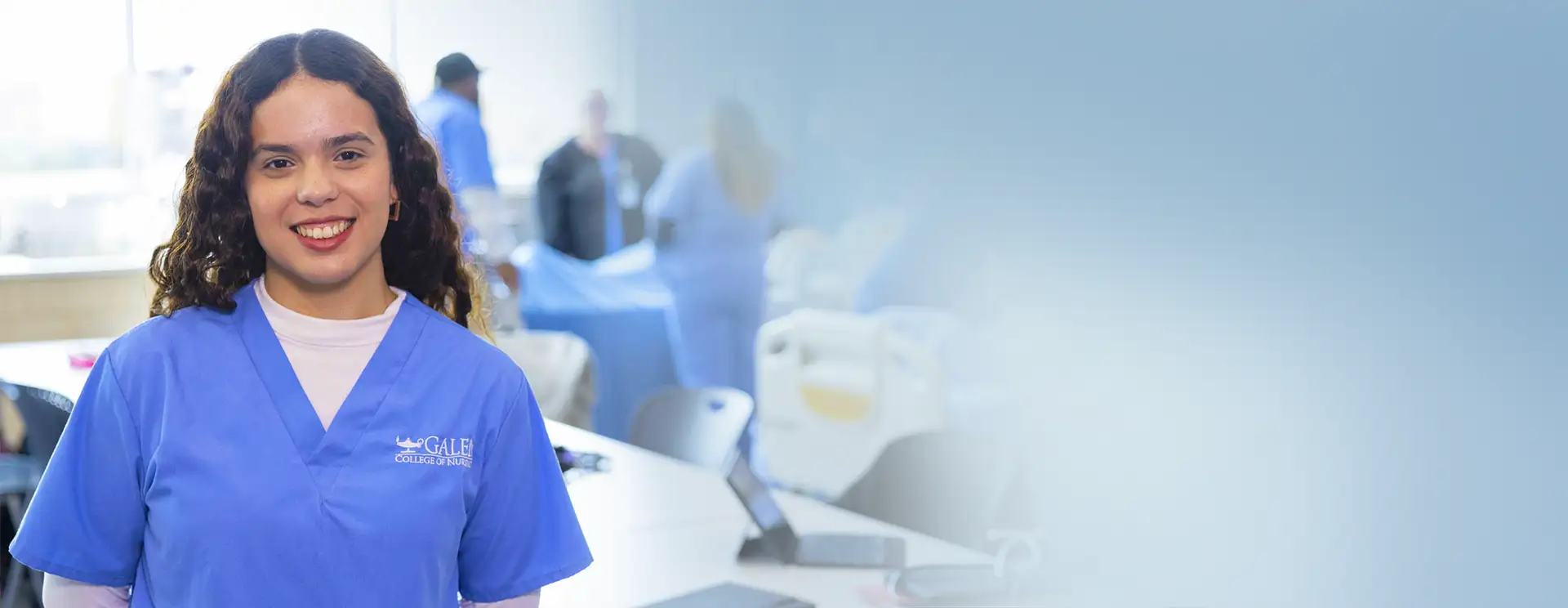 A nursing student in a blue Galen College of Nursing scrub top smiles in the foreground of a clinical classroom. Behind her, several students in scrubs work around hospital beds, while the right side of the image fades to a soft blue gradient.