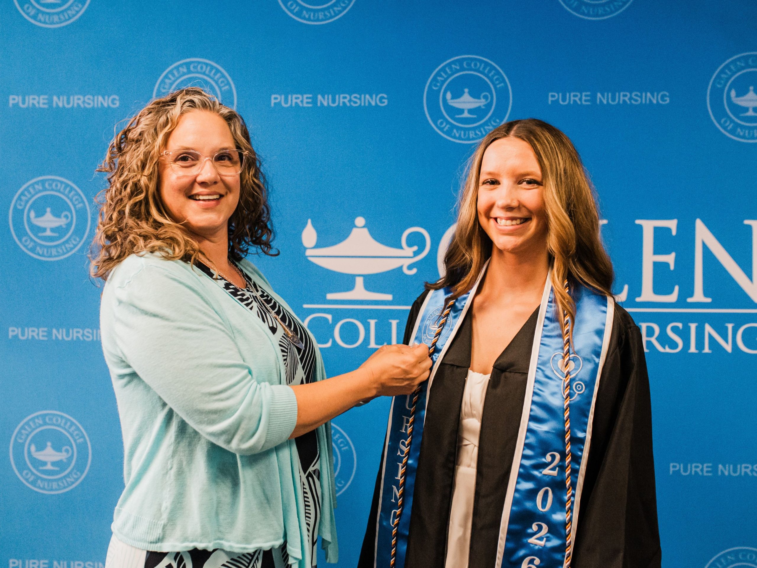 Graduate in cap and gown stands while supporter adjusts nursing stole in front of Galen College of Nursing backdrop.