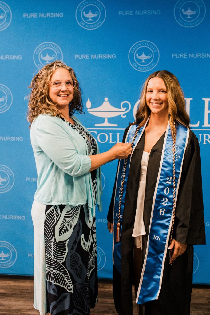 Graduate in cap and gown stands while supporter adjusts nursing stole in front of Galen College of Nursing backdrop.
