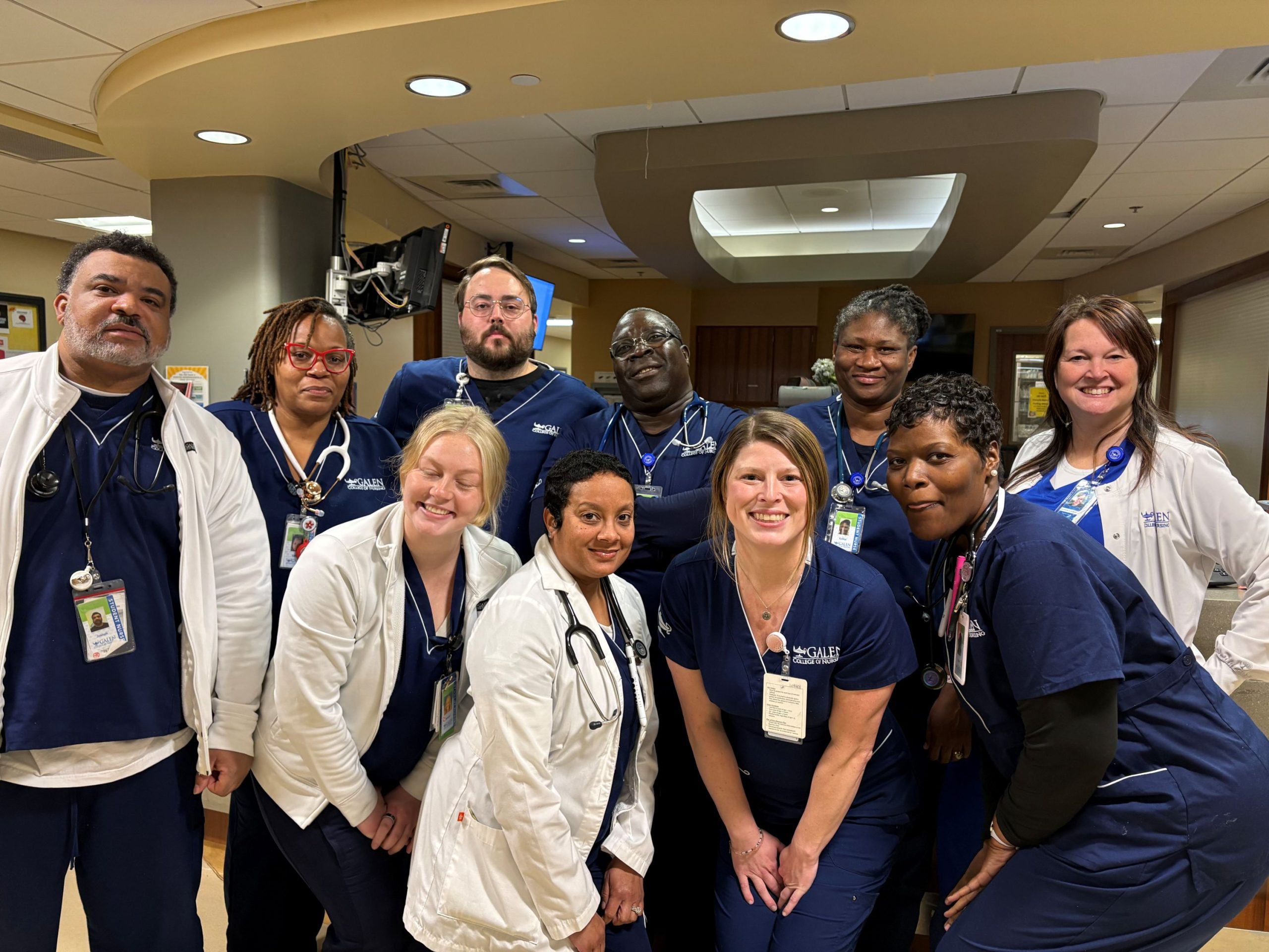 A group of nurses and nursing students wearing navy scrubs and white lab coats pose together in a hospital unit, standing closely as a team in a clinical workspace.