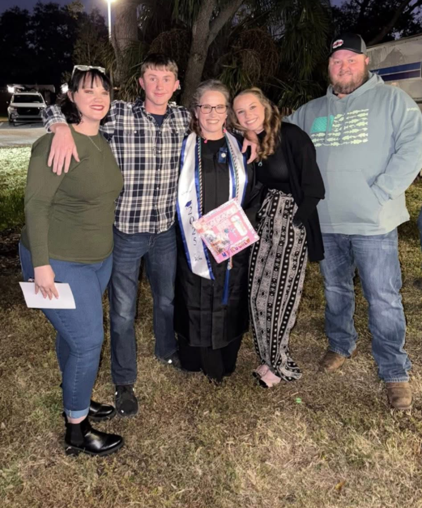 A graduate in cap and gown stands outdoors with family members after a graduation ceremony.