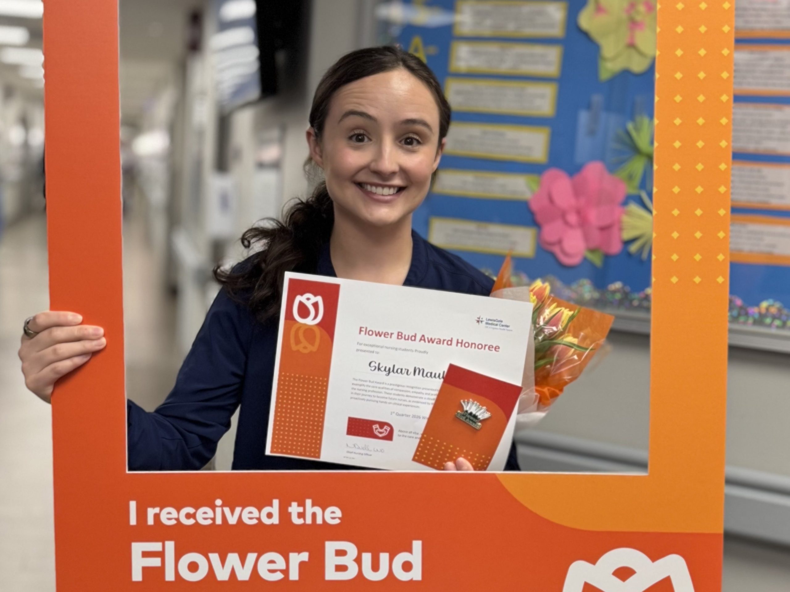 Nursing student poses with a Flower Bud Award certificate and flowers inside a branded photo frame.