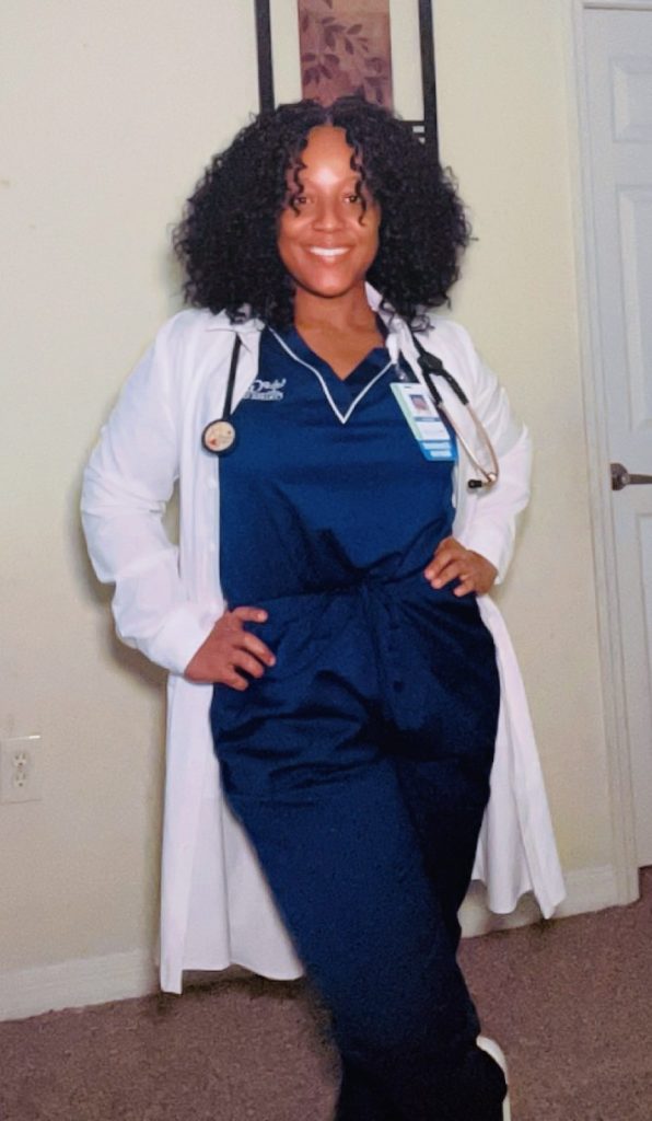 Galen nursing student wearing navy scrubs and a white lab coat, standing confidently indoors with a stethoscope and ID badge.
