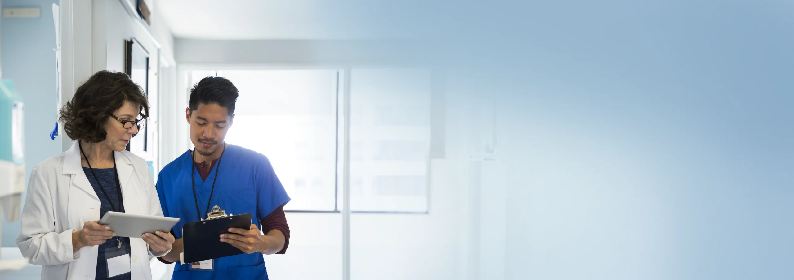 A nursing student in blue scrubs reviews a clipboard while standing beside an instructor in a white lab coat holding a tablet. They appear to be discussing something in a brightly lit hospital hallway that fades into a soft blue gradient on the right.