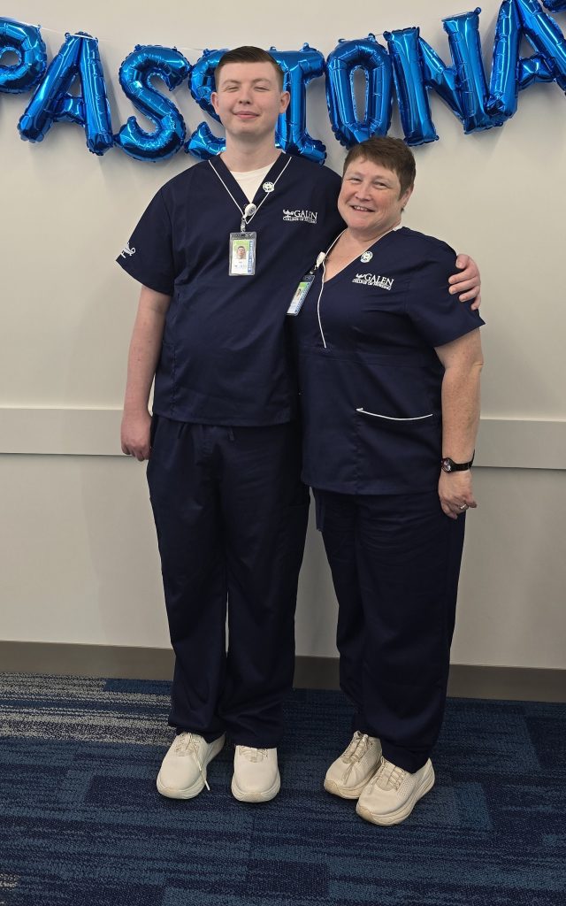 Two Galen College of Nursing students in navy scrubs stand side by side, wearing campus ID badges, with blue balloon letters spelling part of “COMPASSION” behind them.
