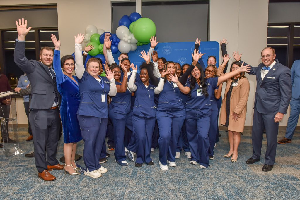 A group of Galen College of Nursing students and campus leaders stand together indoors with raised hands during a celebration, with blue and green balloon décor and a “Pure Nursing” backdrop.