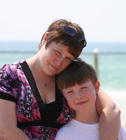 An adult and a child stand close together near a beach, with ocean water and a railing visible in the background.