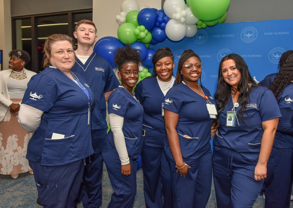 A group of Galen College of Nursing students wearing navy scrubs stand together indoors in front of a blue “Pure Nursing” backdrop with blue, green, and white balloon décor.
