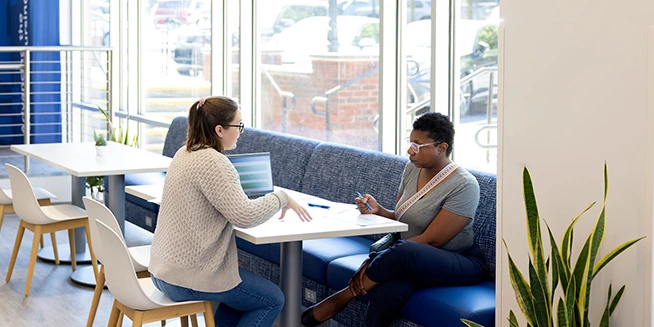 Two women sit at a table in a bright, modern interior with a blue booth, looking at papers and a laptop, possibly discussing something academic.