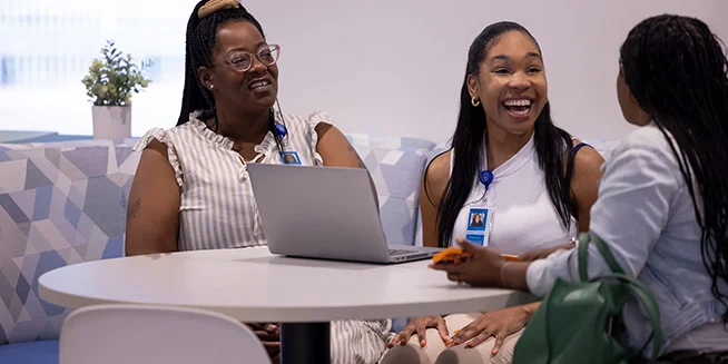 Three women are seated at a round white table, engaged in conversation and smiling. One woman on the left wears a striped top and glasses, looking towards the right. A woman in the middle, wearing a white top, laughs and looks towards the woman on the right, who has her back to the viewer.