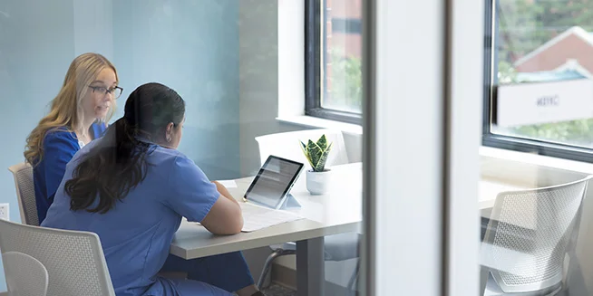 Two women are seated at a white table in a bright room with large windows. One woman, with long dark hair and wearing blue scrubs, has her back to the viewer and is looking at a tablet. Opposite her, a woman with long blonde hair and glasses, wearing a blue top, looks at the tablet as well, collaborating.