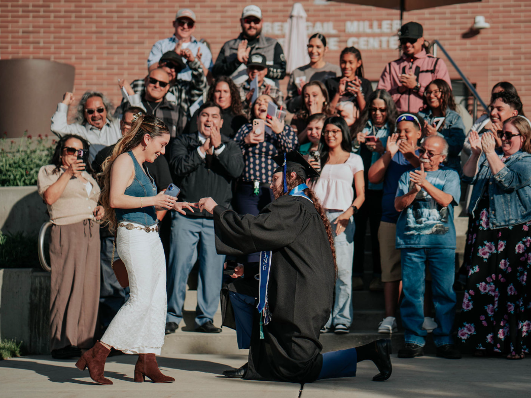 A person in a graduation cap and gown kneels on one knee to place a ring on another person’s hand outdoors, while a large group of people behind them stand on steps clapping and holding phones. A brick building in the background includes partial visible text reading “Central Miller … Center.”
