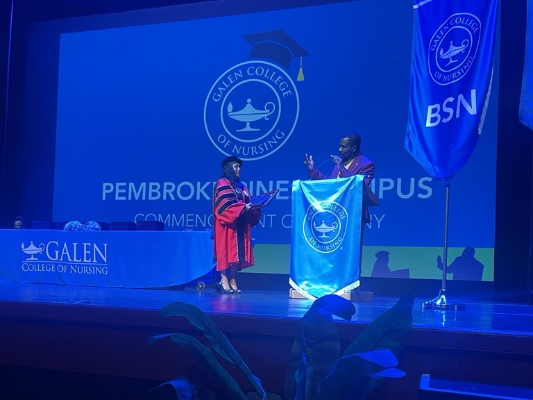 Graduation ceremony stage scene at Galen College of Nursing’s Pembroke Pines campus. One person in academic regalia stands on stage near a podium decorated with a Galen banner, while another person stands nearby. A large screen behind displays the college logo, and a vertical banner labeled “BSN” is visible to the right. A potted plant sits in the foreground near the stage.