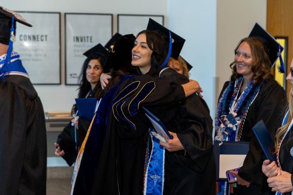 Graduates in caps and gowns gather indoors as one graduate embraces another while holding a diploma, with fellow graduates standing nearby holding certificates.