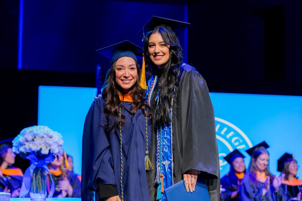 Two graduates in caps and gowns stand together on stage during a commencement ceremony, holding diplomas with a lit stage and seated faculty in the background.